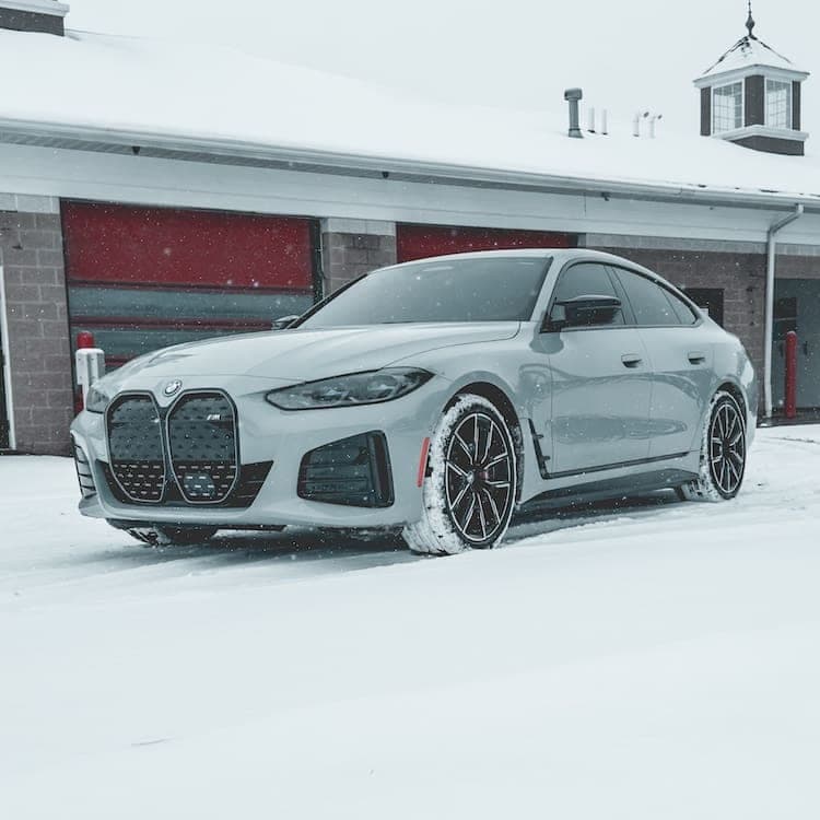 The light grey BMW i4 M50 parked in snow outside a building with red garage doors and a rooftop cupola.