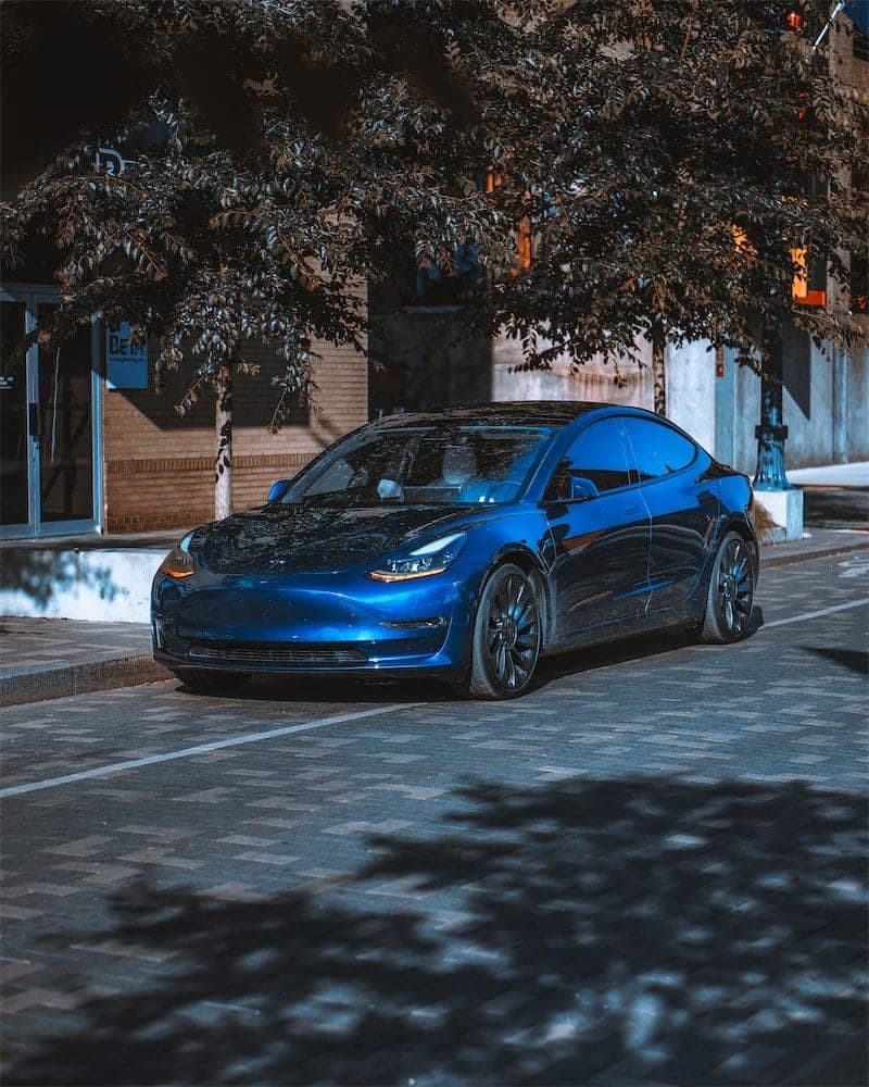 The blue Model 3 parked on a brick street under a tree, front three-quarter view.