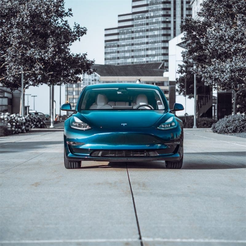 The blue Model 3 head-on across a concrete plaza, trees and a tall building behind.