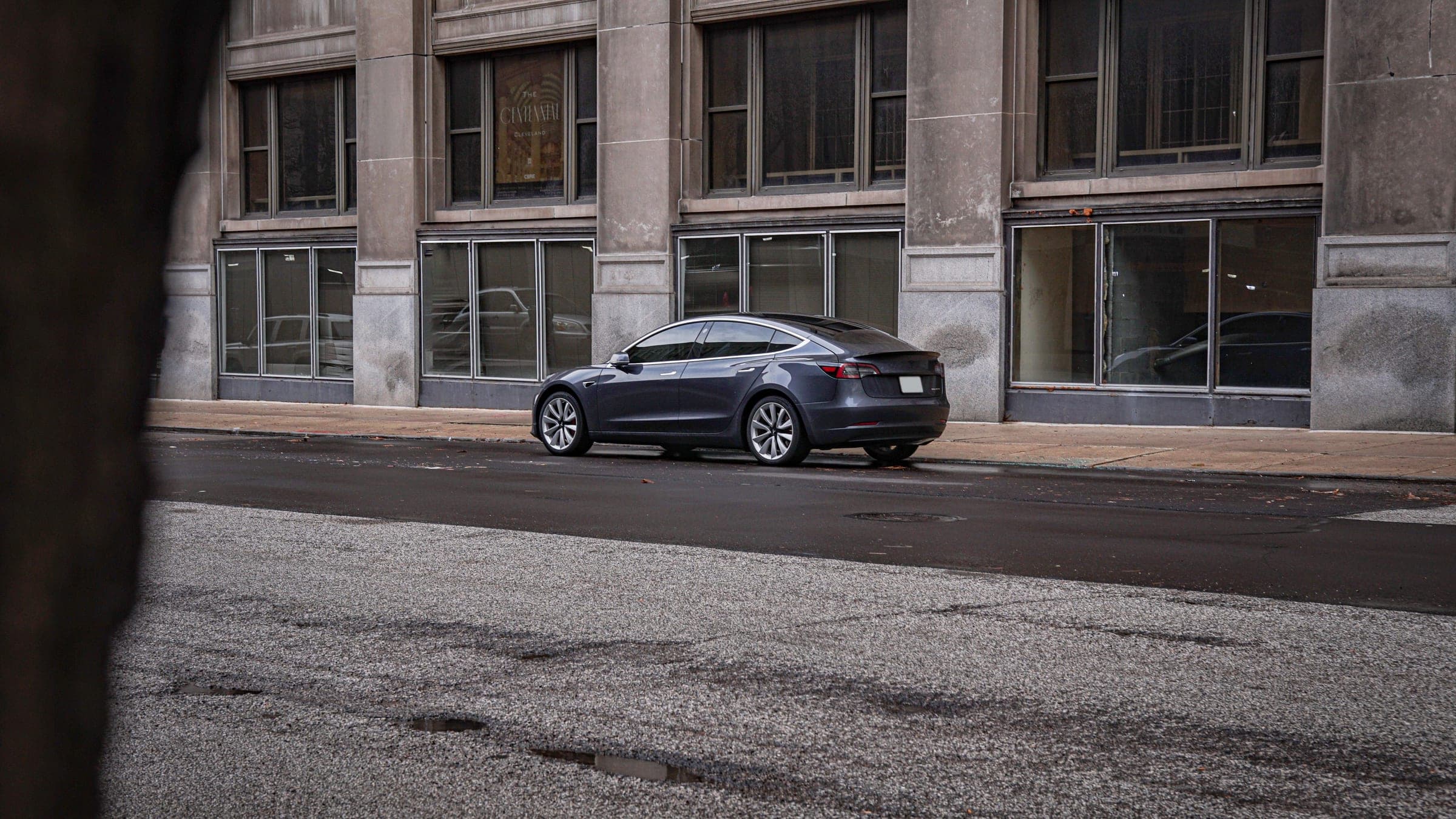 A grey Model 3 parked across the street from a stone office building.